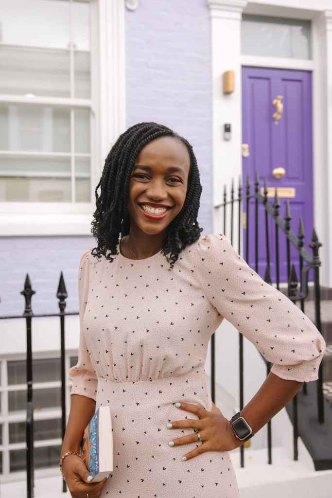 afoma umesi stands in front of purple door with hand on waist holding a book